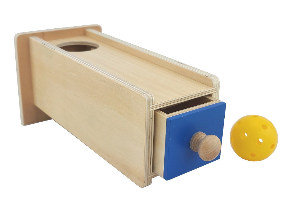 Object permanence box with blue drawer and hollow yellow plastic ball. Box has a circular hole for the ball to be dropped into.