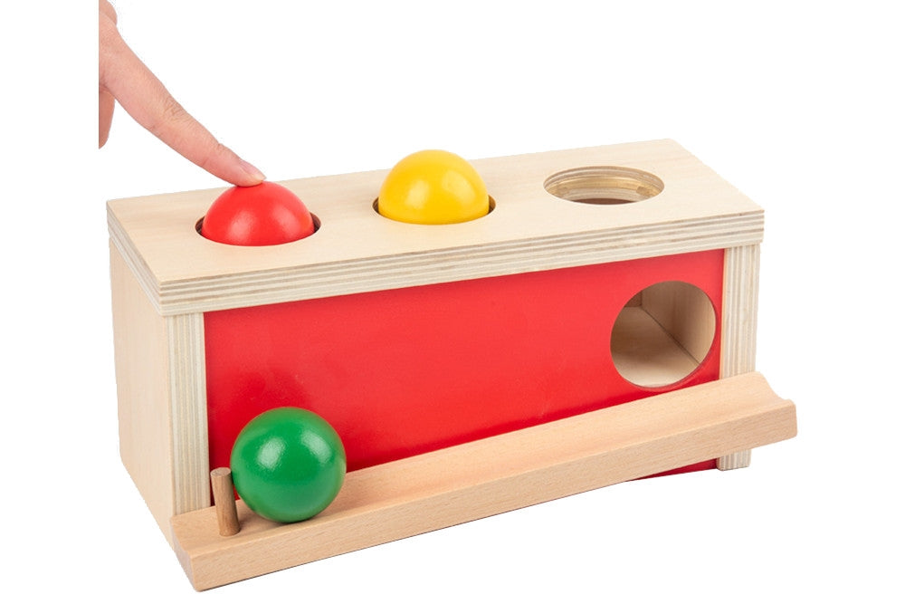Natural wood ball push toy with red side and three balls in red, yellow, and green. A finger demonstrates pushing red ball through hole.