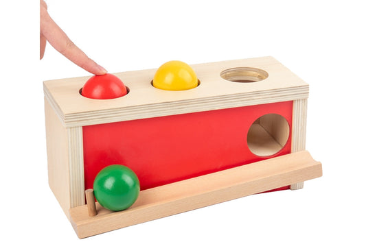 Natural wood ball push toy with red side and three balls in red, yellow, and green. A finger demonstrates pushing red ball through hole.