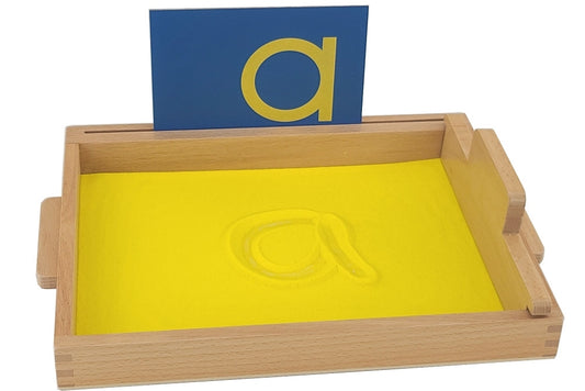 Sand tracing tray filled with yellow sand. The letter 'a' is traced into the sand, and the corresponding blue sandpaper letter is inserted into the slot.